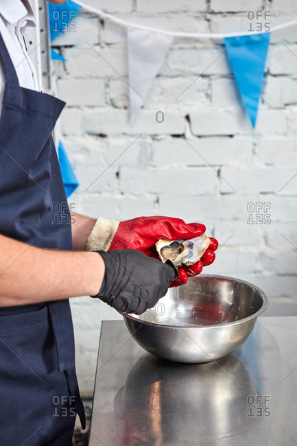 A man in a black apron opens a shell of oysters on a metal surface around a white brick wall. Cooking gourmet health food