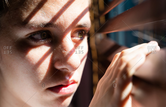 Youth girl with green eyes opening a wooden curtain with her fingers to look through the window