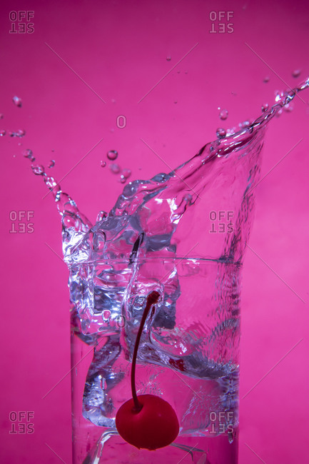 Splashing cocktail with ice and cherry on pink background
