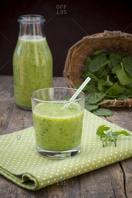 Glass bottle and glass of green smoothie- made of spinach- rocket salad- apple- orange- banana and cucumber- on cloth wooden table
