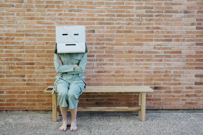 Woman sitting on bench in front of brick wall with serious  face on cardboard box- arms crossed