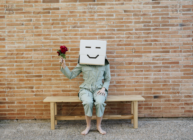 Woman holding red rose- wearing cardboard box with happy face- sitting on bench in front of brick wall