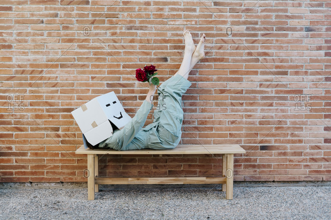 Woman holding red rose- wearing cardboard box with happy face- lying on bench in front of brick wall