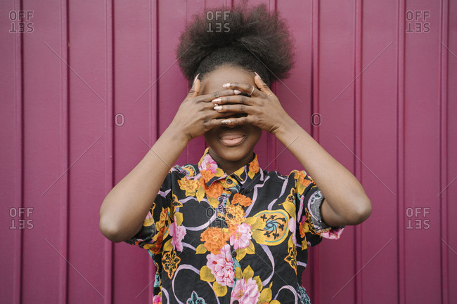 Smiling young woman weaning blouse with floral design covering eyes with her hands