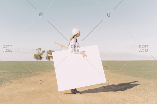 Young woman carrying easel and empty canvas on dry field