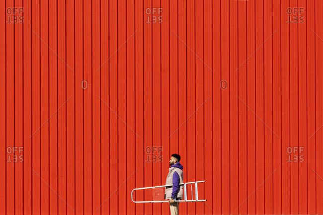 Young man carrying a ladder in front of a red wall
