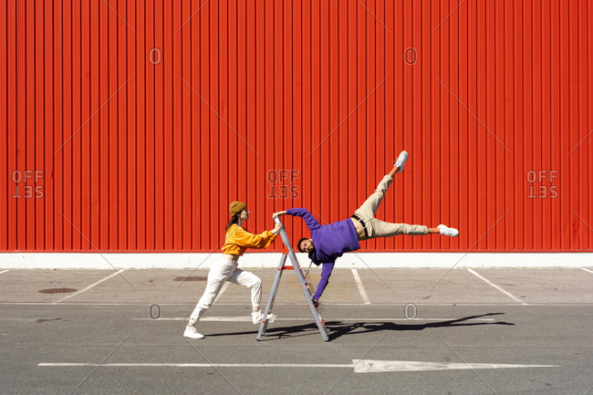 Young man and woman performing with a ladder in front of a red wall