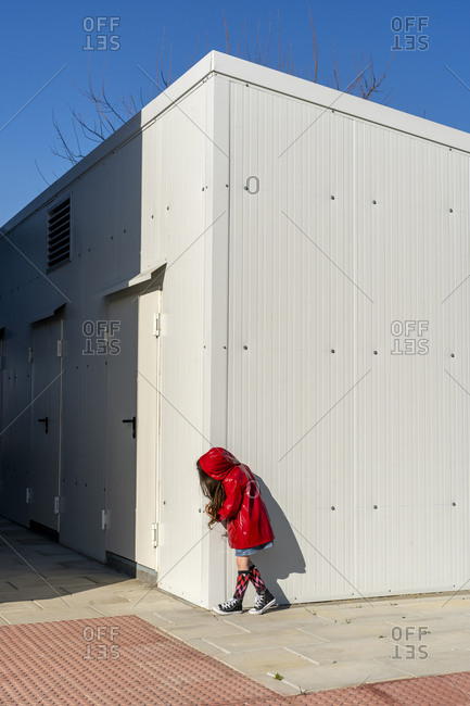 Little girl wearing red rain coat hiding behind a corner