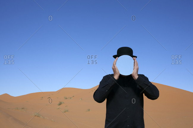 Morocco- Merzouga- Erg Chebbi- man wearing a bowler hat holding mirror in front of his face in desert dune