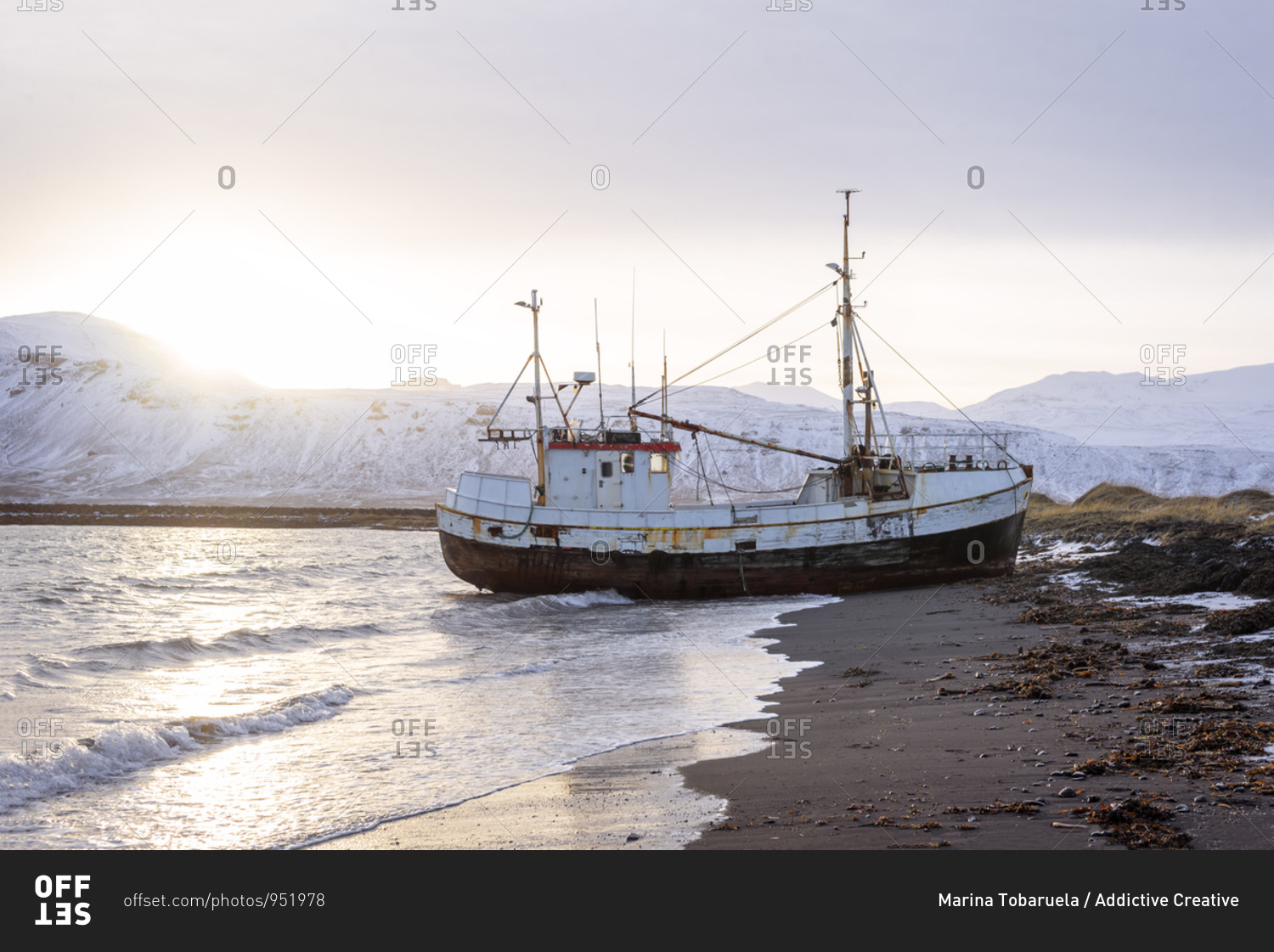 Distant old boat washed by sea surf stranding on shore against backdrop of snowy mountain ranges and beautiful sunset in winter in Iceland