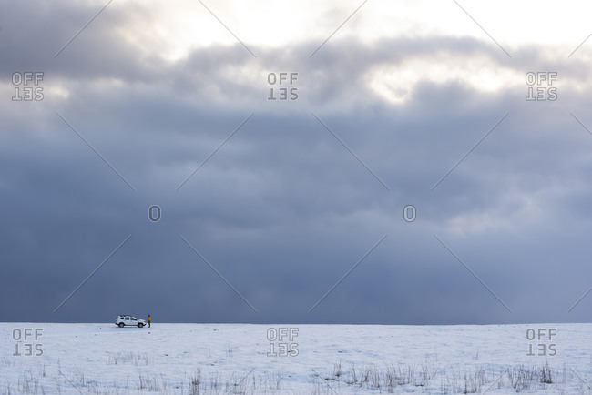 Cold winter landscape with snowy white terrain with white car and remote lonely traveler against backdrop of endless delightful cloudy sky in cold winter
