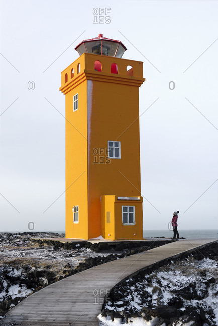 Beautiful large lighthouse with yellow walls and red roof locating on rocky shore near sea water and distant tourist standing near on background of cloudy sky and gray cold sea