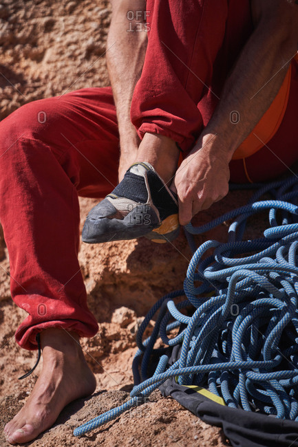 Unrecognizable male alpinist in red sportswear sitting on rocky ground near blue rope and wearing climbing shoes before climbing