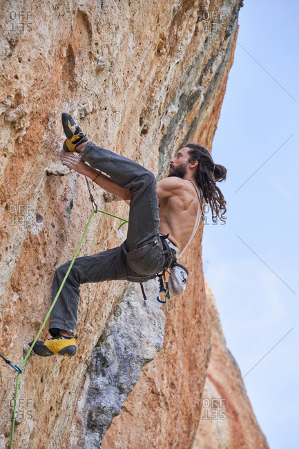 From below side view of shirtless bearded alpinist in safety equipment climbing up on rocky slope of mountain and looking up