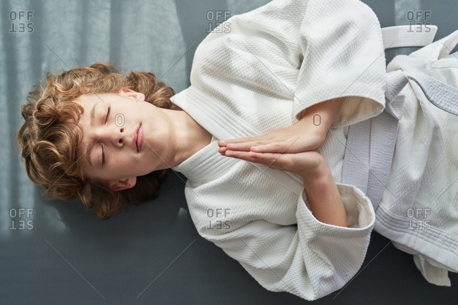 Thoughtful young boy in white kimono lying down with hands together while practicing meditation with closed eyes during training judo at home