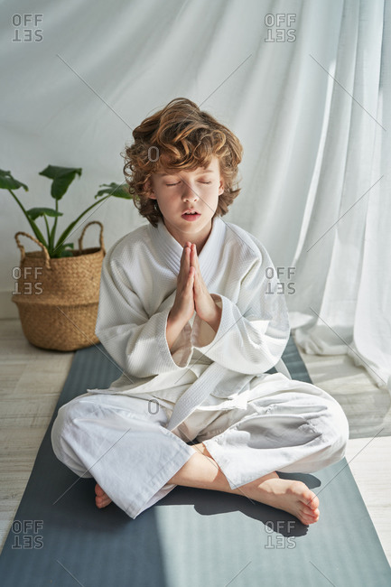 Thoughtful young boy in white kimono sitting with legs crossed and hands together while practicing meditation with closed eyes during training judo at home