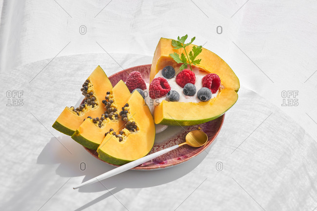 From above pieces of ripe papaya with yoghurt and fresh berries placed near spoon on plate on white background