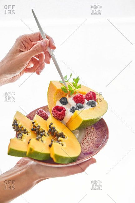 Unrecognizable person carrying plate with fruit dessert and poking slices of papaya with yoghurt and berries with stick against white background