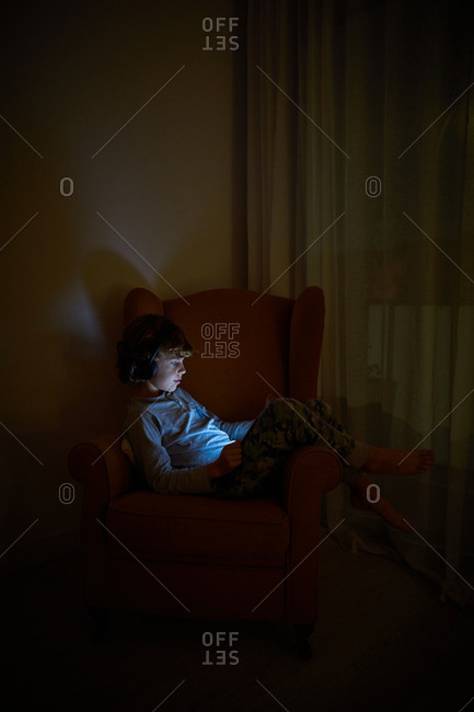 Stock vertical photo of a child sitting on an armchair in a corner of a room looking at tablet computer with headphones at night. Technology