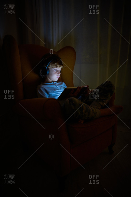 Stock vertical photo of a child sitting on an armchair in a corner of a room with a tablet in his lap with headphones at night. Technology