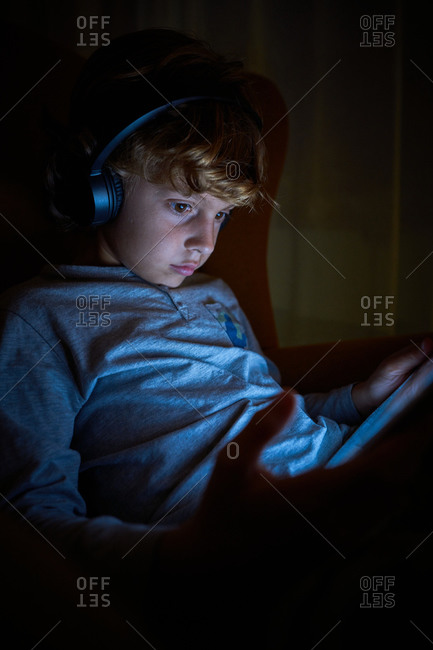 Stock vertical photo of a child sitting with headphones on a sofa with a tablet at night. Technology