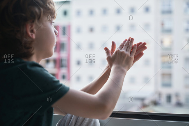 Side view of crop tranquil preteen boy looking out window with interest to yard against multistory building and clapping hands while sitting on window sill in light room