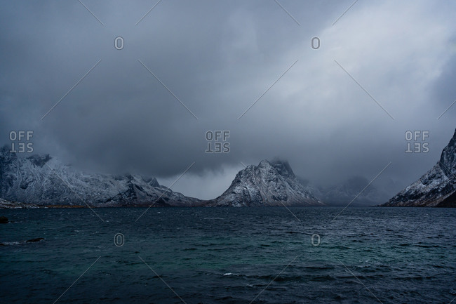 Breathtaking view of blue rippled sea water against snowy mountain ridges on shore under gray cloudy sky in wintertime in Norway