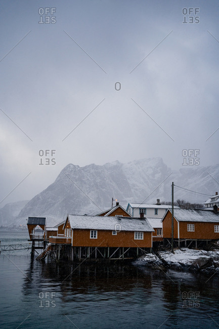 Yellow country houses on coast of strait against misty snowy mountain crests in overcast weather in Norway