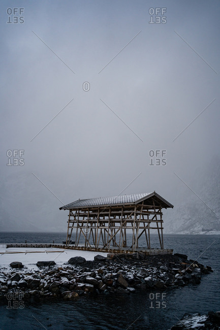 Solitary construction with wooden pillars and gray roof on stone beach washing by troubled water against misty mountain ridges in overcast weather in harbor in Norway