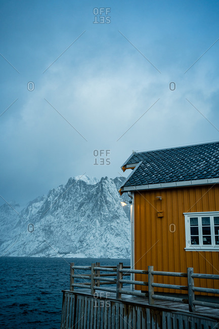 Yellow country houses on coast of strait against misty snowy mountain crests in overcast weather in Norway