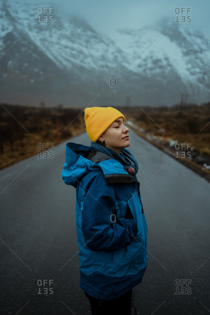 Side view of happy relaxed woman in blue warm clothes and bright yellow hat enjoying life while standing with eyes closed on asphalt road going to snowy foggy mountains in Lofoten