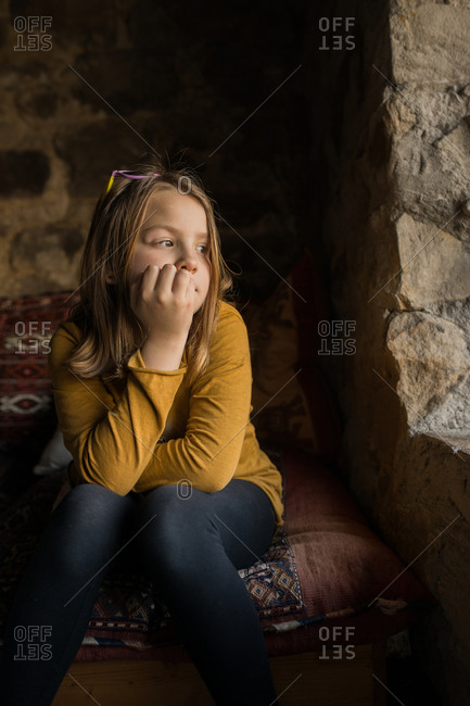 Lonely thoughtful girl in casual clothes leaning on hand while sitting on couch and looking away in window of old stone house in Spain