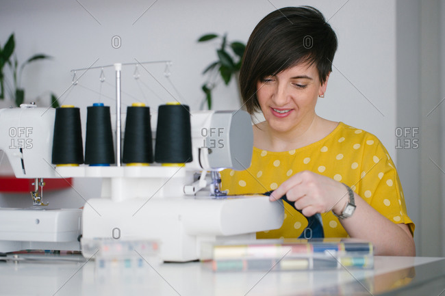 Happy brunette adult woman smiling and using sewing machine to make denim garment while working in home workshop