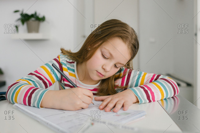 Focused girl in striped t shirt writing in notebook while sitting at table and doing homework at home
