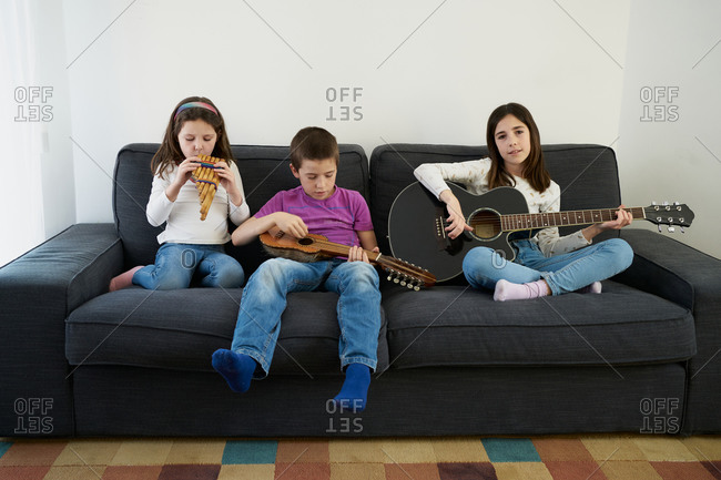 Positive young little boy and girls in casual clothes sitting on comfortable sofa together and playing music instruments while spending time together at home