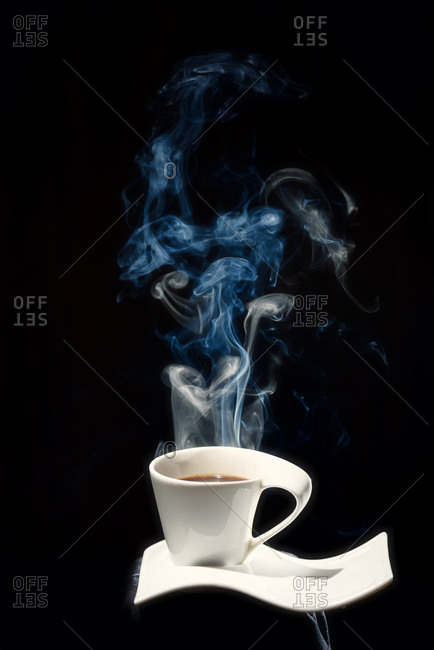 White ceramic cup of fresh hot coffee with steam rising up placed on white ceramic plate in dark room on black background