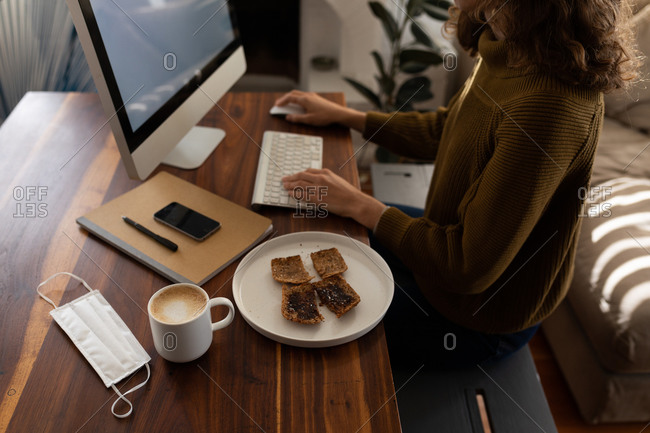 Mid section of a Caucasian woman spending time at home, sitting by her desk and working using her computer, with a snack and coffee put next to her. Social distancing and self isolation in quarantine lockdown.