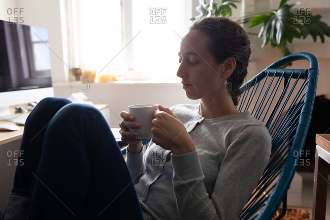 Caucasian woman spending time at home, sitting on a chair and holding a cup of coffee, relaxing. Social distancing and self isolation in quarantine lockdown.