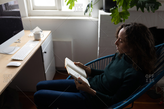 Caucasian woman spending time at home, sitting on a chair and reading a book, relaxing. Social distancing and self isolation in quarantine lockdown.