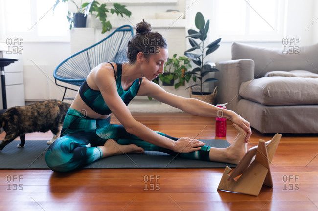 Caucasian woman spending time at home, wearing sportswear, sitting on a yoga mat and stretching up, joining online yoga course, using her tablet. Social distancing and self isolation in quarantine lockdown.