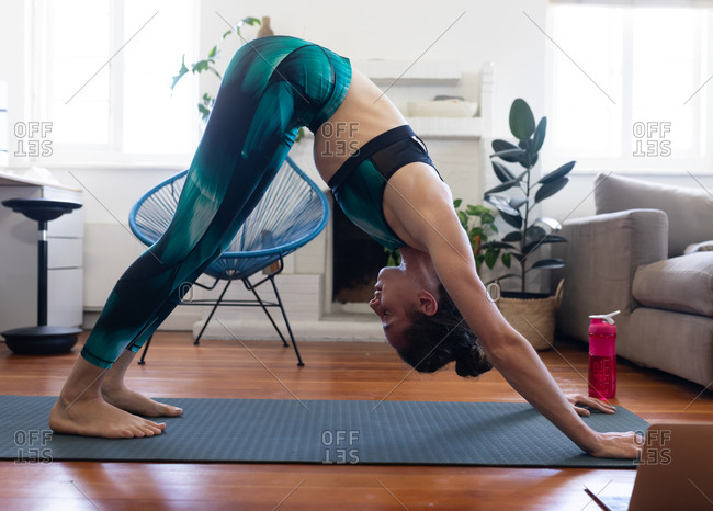 Caucasian woman spending time at home, wearing sportswear, sitting on a yoga mat and stretching up, joining online yoga course, using her laptop. Social distancing and self isolation in quarantine lockdown.