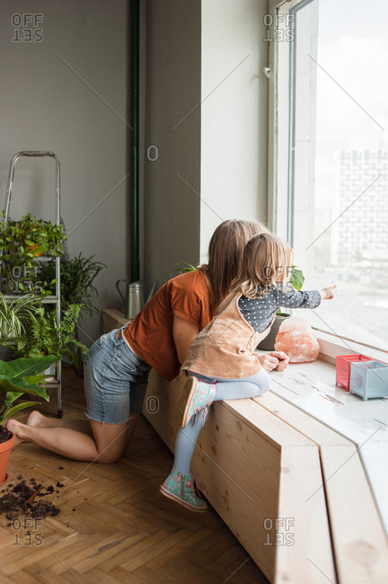 Mother and daughter look outside kneeling at window. Kid points.
