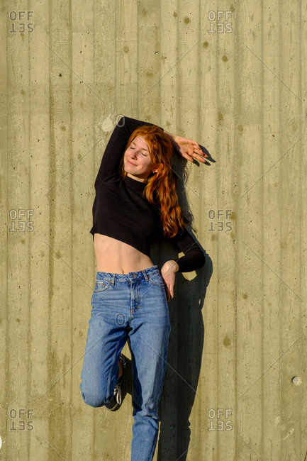 Portrait of smiling teenage girl with eyes closed leaning against concrete wall enjoying sunlight