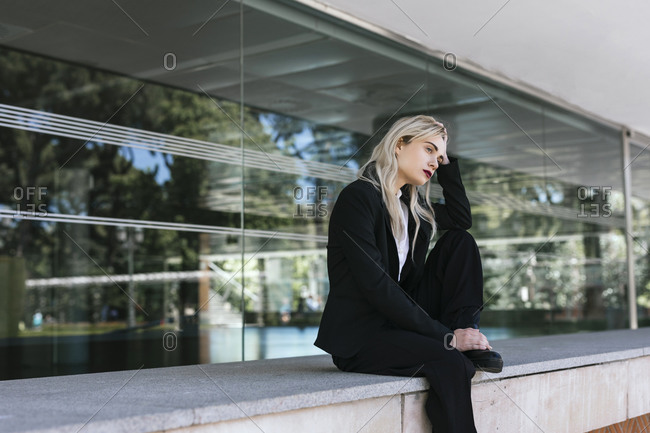 Pensive young businesswoman with dyed blond hair sitting in front of office building looking at distance
