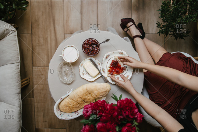 Elegant woman in high heels holding a piece of bread with raspberry jam and butter by fresh peonies