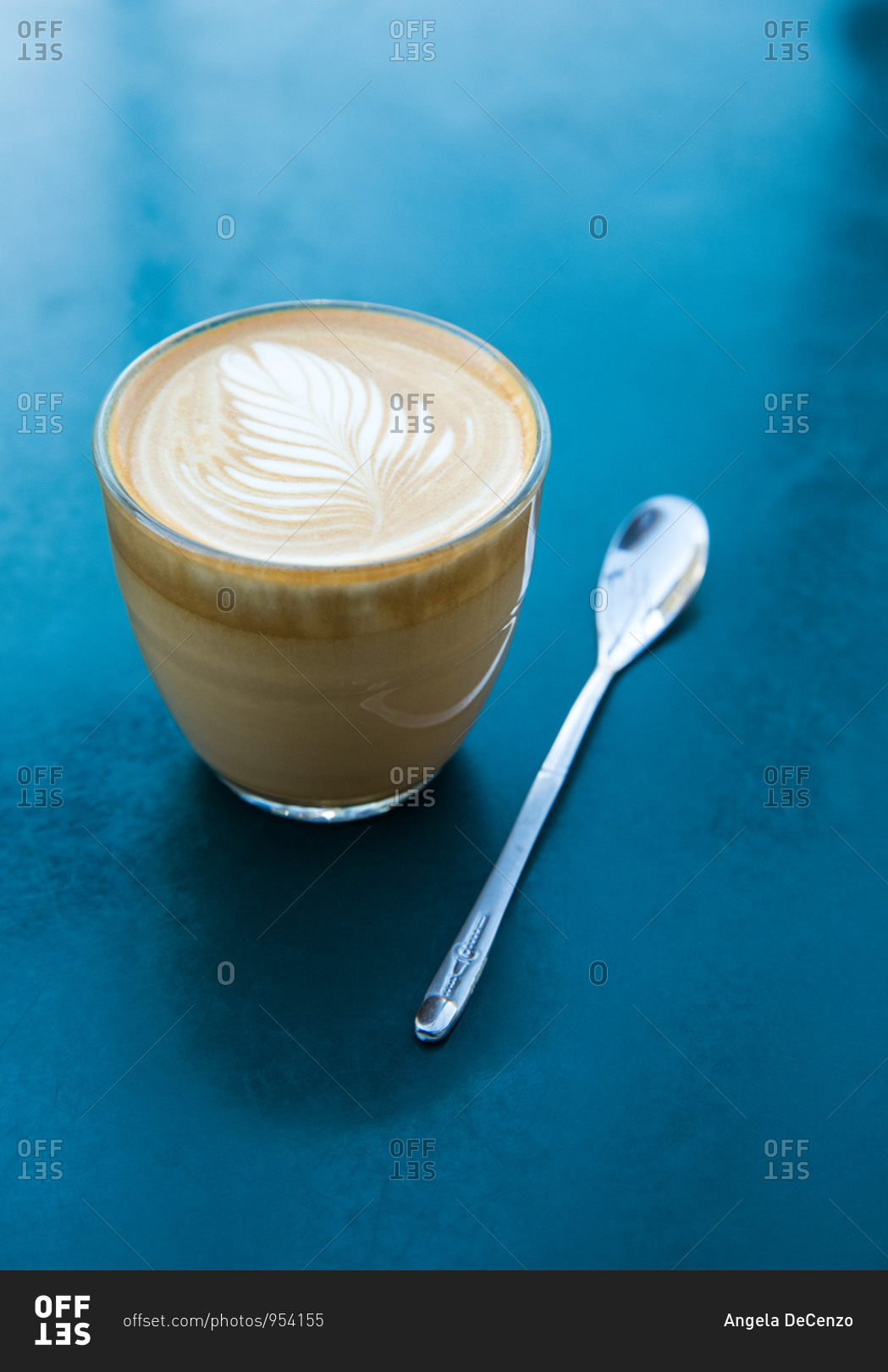 A cortado and small spoon sitting on a cerulean blue surface