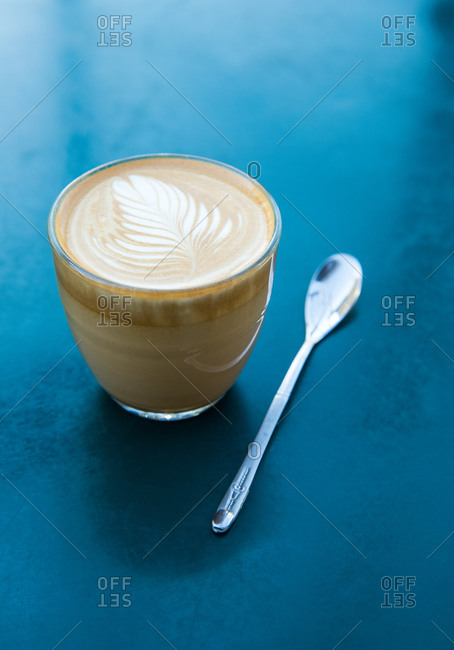 A cortado and small spoon sitting on a cerulean blue surface