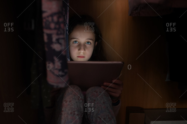 Little girl in sleepwear sitting in dark room with glowing tablet