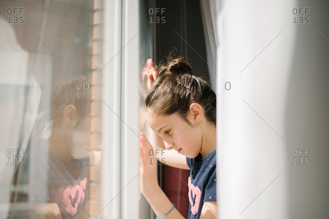 Side view of sad girl standing near window with hands on glass and looking in bright sunlight missing friends