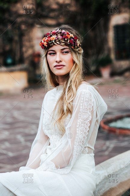 Pretty young woman in translucent white dress and floral wreath looking away while sitting on blurred background of yard on wedding day